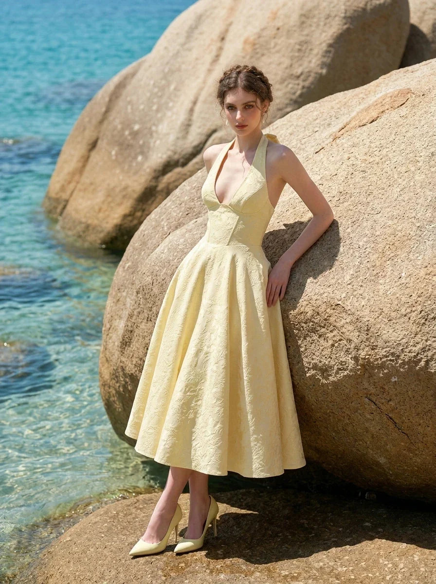 Woman posing next to a large rock on a beach, wearing a pale yellow jacquard halter neck midi dress.