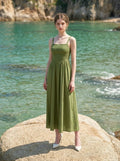 Woman wearing a green linen blend midi dress with a smocked bodice and spaghetti straps, standing on a rock with turquoise water in the background.