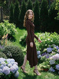 Model wearing a long-sleeved brown ruched mini dress with an asymmetrical hemline and high neckline, walking in a garden