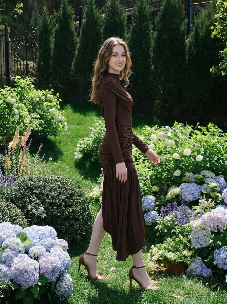Model wearing a long-sleeved brown ruched mini dress with an asymmetrical hemline and high neckline, walking in a garden