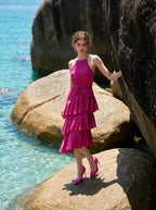 Woman wearing a fuchsia satin halter neck tiered midi dress with a self-tie belt, posing outdoors by the ocean