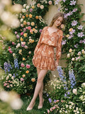 Woman wearing a floral print mini dress with long sleeves and a tie-neck leaning against a wall with flowers