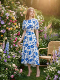 Woman wearing a white and blue floral print midi dress with puff sleeves and a cut-out waist, posing in a garden.