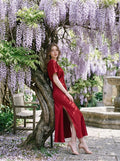 Woman in a red satin V-neck dress with kimono sleeves and a high slit, leaning against a tree with wisteria