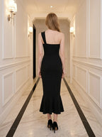 Back view of a woman in a black one-shoulder mermaid dress with a sweetheart neckline and ruched bodice, standing in a hallway.