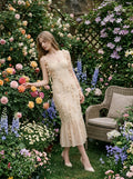 Woman wearing a cream-colored midi dress with 3D floral embellishments and a ruffled hem, standing in a garden with roses and a chair.