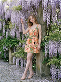 Woman in a peachy-pink, fruit and coffee cup print dress with a scoop neck and sleeveless design, standing near a wisteria-covered wall.