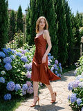 Woman walking in a rust-colored satin slip dress with spaghetti straps, a cowl neckline, and an asymmetrical hemline, in an outdoor garden setting with blue hydrangeas.