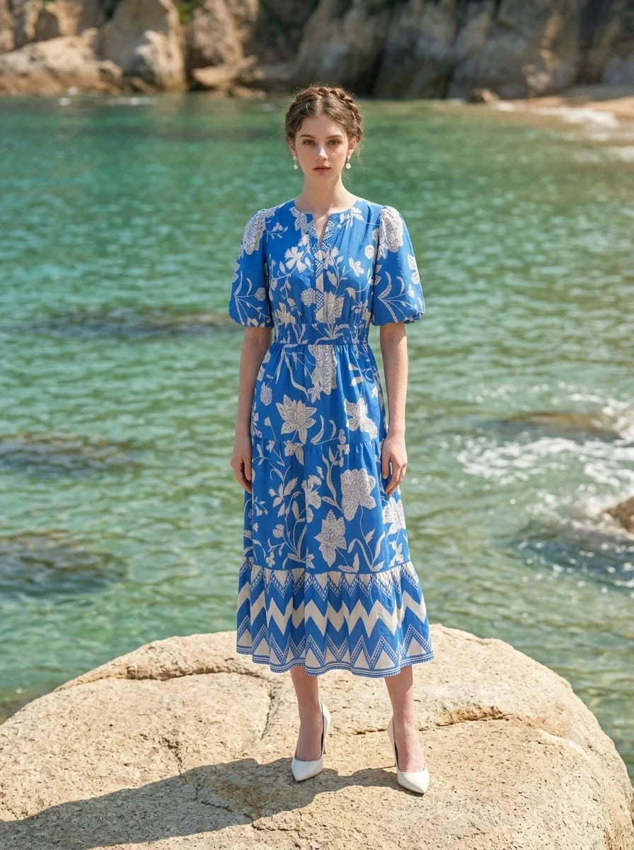 Woman wearing a blue floral print midi dress with puff sleeves, standing on a rock formation by the ocean.