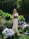 Woman in a cream satin maxi dress with black lace detailing at the waist and a halter neckline, walking through a garden with hydrangeas.