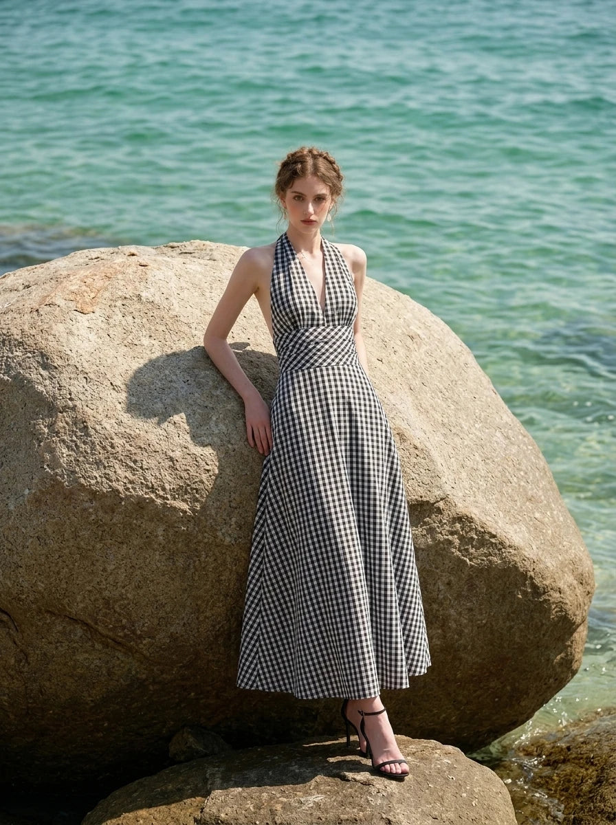 Woman leaning against a large rock on a beach, wearing a black and white gingham check halter neck maxi dress with a plunging V-neck and a defined empire waist.