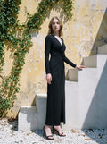 Woman in a black long-sleeved V-neck gown with a front knot and high slit, standing on outdoor steps with a yellow wall in the background.