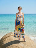 Woman standing on a large rock at the beach wearing a white midi dress with a colorful abstract graphic print and gold chain spaghetti straps.