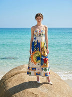 Woman standing on a large rock at the beach wearing a white midi dress with a colorful abstract graphic print and gold chain spaghetti straps.