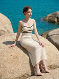 Woman wearing a cream knit maxi dress with black striped bodice, sitting on a rock by the sea, profile view