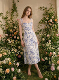 Woman wearing a white midi dress with a blue floral print and tie shoulder straps, posing in a floral garden.