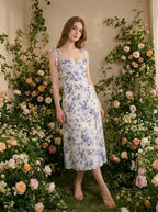 Woman wearing a white midi dress with a blue floral print and tie shoulder straps, posing in a floral garden.