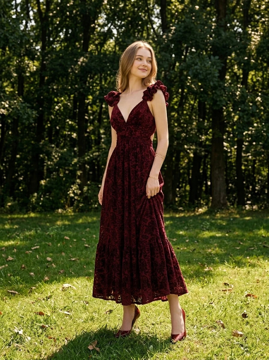 Woman in a burgundy velvet burnout A-line maxi dress with a V-neckline and ruffled cap sleeves, standing in a grassy area with trees in the background.