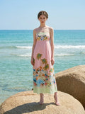 Woman standing on a large rock by the ocean, wearing a pink midi dress with spaghetti straps and a tropical print.
