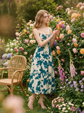 Woman in a cream and teal floral print midi dress with spaghetti straps and a tie-front bodice, interacting with flowers in a garden setting.