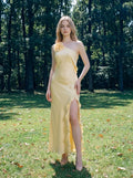 Woman in a pale yellow satin one-shoulder gown with a ruffled high slit in a grassy, wooded area.