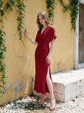Woman in a red satin V-neck dress with kimono sleeves and a high slit, standing by a yellow wall