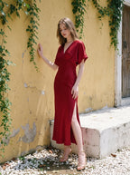 Woman in a red satin V-neck dress with kimono sleeves and a high slit, standing by a yellow wall