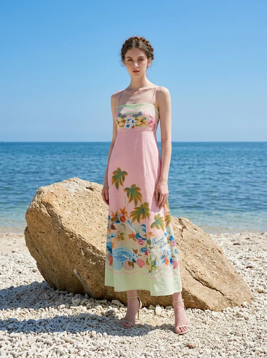 Woman standing on a beach with large rocks, wearing a pink midi dress with spaghetti straps and a tropical print.