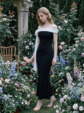 Woman in a black off-the-shoulder mermaid dress with a white draped accent on the shoulder, posing in a lush garden with flowers.