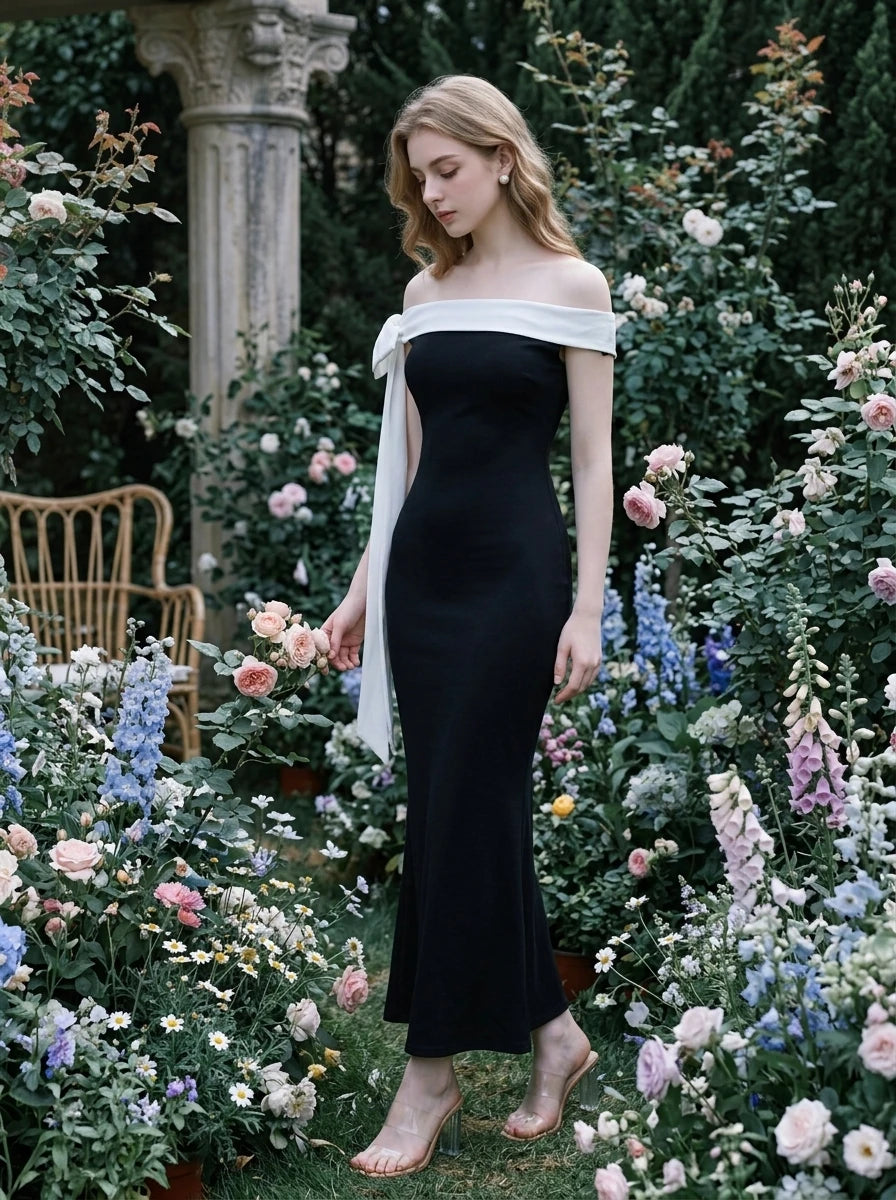 Woman in a black off-the-shoulder mermaid dress with a white draped accent on the shoulder, posing in a lush garden with flowers.