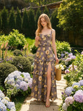 Woman wearing a grey floral print maxi dress with ruffles and a high slit, holding a straw bag in a garden.