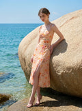 Woman in a peach floral print midi dress with side slit posing by a large rock near the ocean