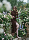 Model wearing a long-sleeved brown ruched mini dress with an asymmetrical hemline and high neckline, leaning against a stone wall with flowers