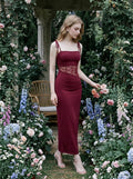 Woman standing in a garden wearing a burgundy dress with a lace inset and square neckline.