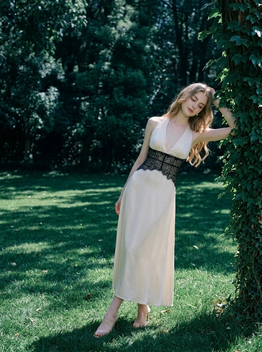 Woman in a cream satin maxi dress with black lace detailing at the waist and a halter neckline, leaning against a tree in a garden.