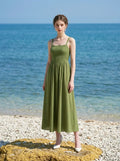 Woman wearing a green linen blend midi dress with a smocked bodice and spaghetti straps, standing on a rock by the sea.