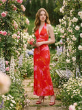 Woman wearing a long red halter neck dress with a tropical leaf print, holding a bouquet of flowers in a garden.