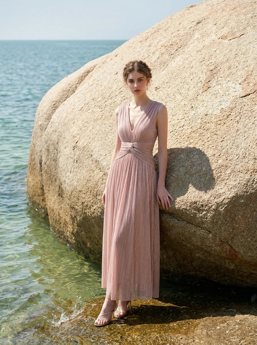 woman in a rose gold metallic a-line maxi dress with a v-neckline and ruched empire waist standing by a rock at the beach