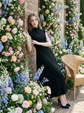 Woman in a sleeveless black A-line midi dress posing against a brick wall with floral decorations.