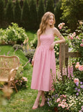 Woman wearing a pink midi dress with spaghetti straps and a square neckline, leaning on a wooden fence in a garden.