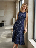 Woman leaning against a wall in a navy blue polka dot midi dress holding a coffee cup