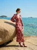 Woman leaning against a large rock by the sea wearing a long, floral print, V-neck dress with long balloon sleeves.