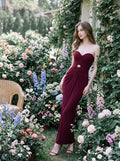 Woman wearing a floor-length, strapless burgundy gown with ruching and gold embellishment, leaning against a wall in a garden with flowers.
