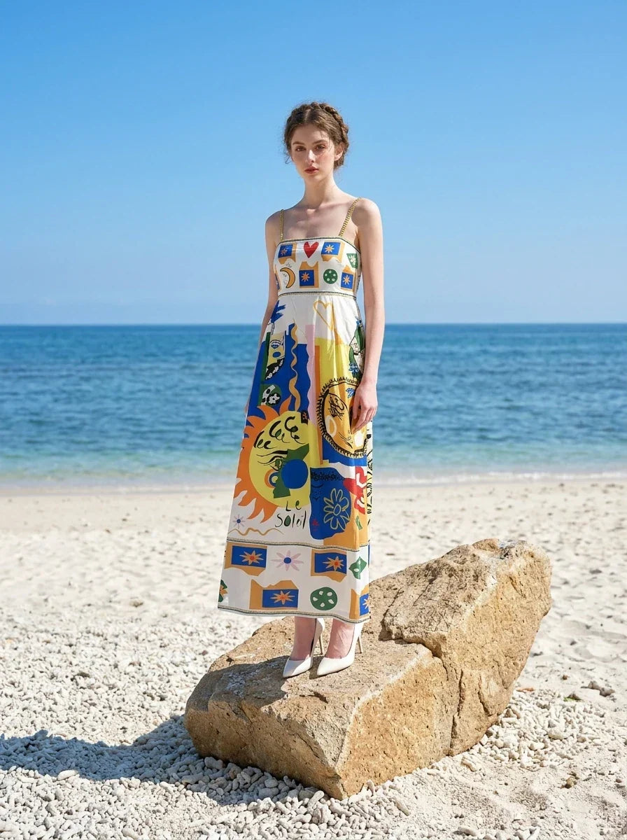 Woman standing on a rock on a beach wearing a white midi dress with a colorful abstract graphic print and gold chain spaghetti straps.