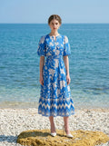 Woman wearing a blue floral print midi dress with puff sleeves, standing on a pebble beach with the ocean in the background.