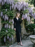 Woman in a black long-sleeved V-neck gown with a front knot and high slit, posing near a stone wall covered in purple wisteria.
