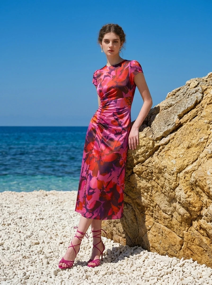 Woman leaning against a rock on a beach, wearing a pink and red floral print satin ruched midi dress.