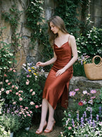 Woman sitting in a rust-colored satin slip dress with spaghetti straps and a cowl neckline, in an outdoor garden setting with pink and purple flowers.