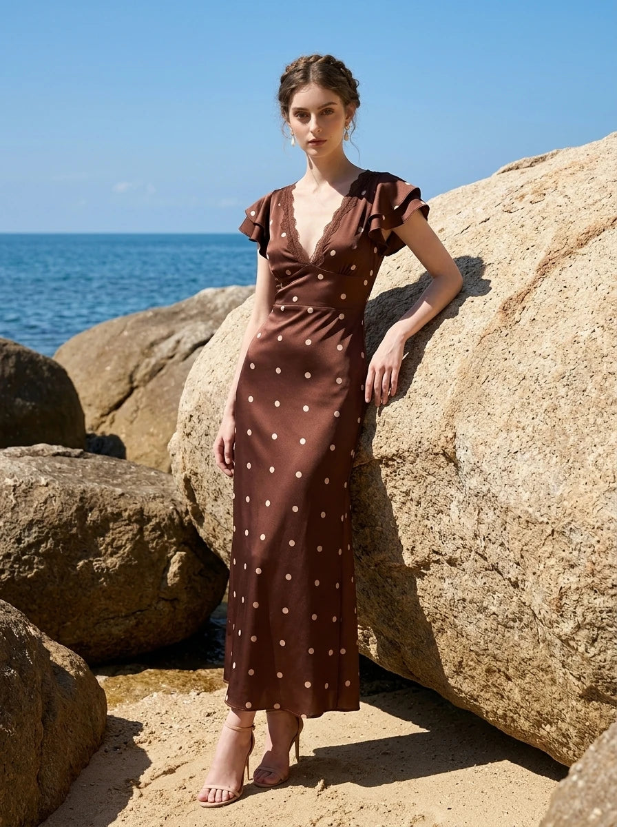 Woman in a brown polka dot midi dress with ruffled sleeves standing by rocks near the ocean.