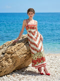 Woman leaning against a large rock on a beach, wearing a white maxi dress with a red floral print and red trim.